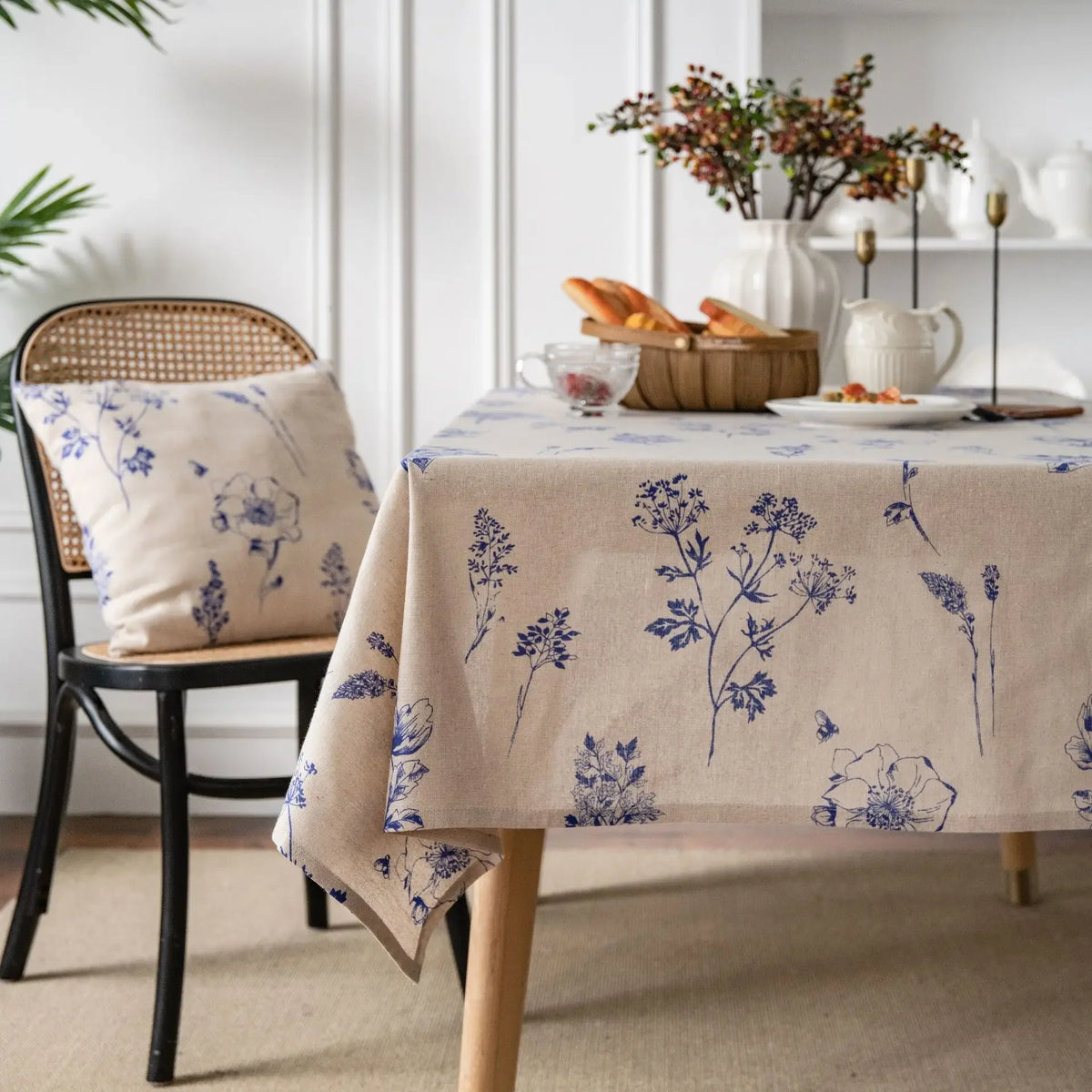 A linen tablecloth with a blue botanical print is displayed on a wooden table in a bright room.