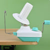 A white hand-operated wool yarn winder device with a clear plastic spool and a metal handle is shown on a wooden table.