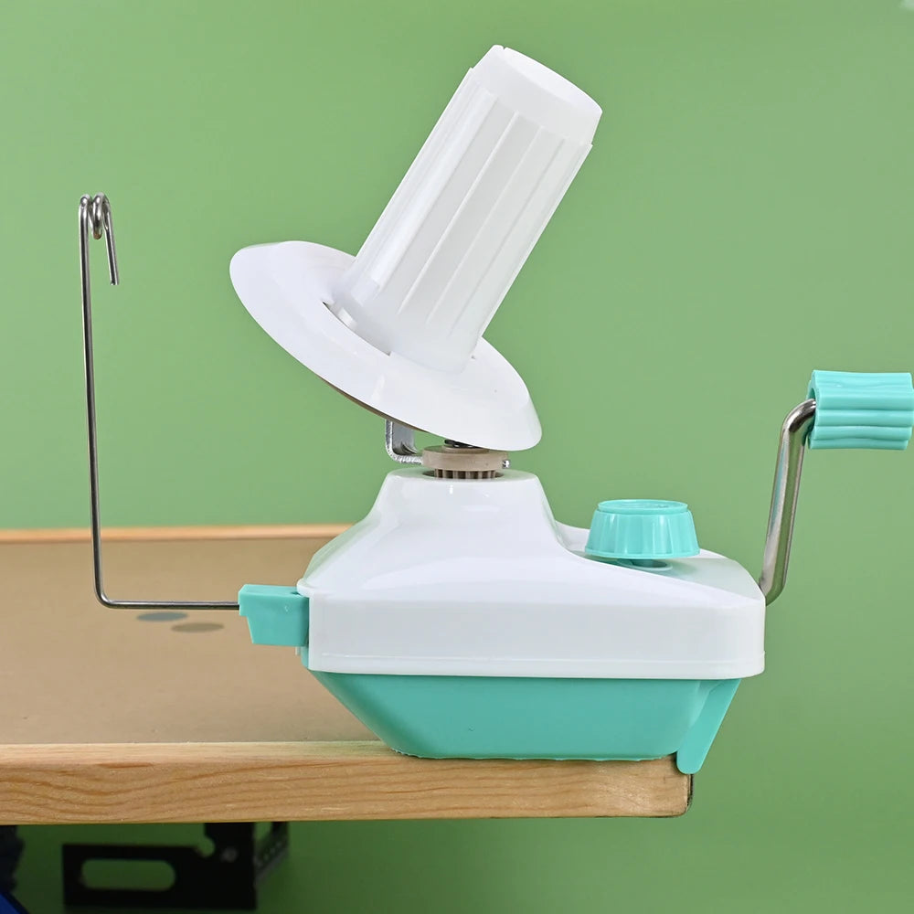 A white hand-operated wool yarn winder device with a clear plastic spool and a metal handle is shown on a wooden table.