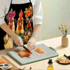 A person wearing a black apron with a funny chicken and floral print is preparing salmon on a cutting board.