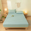 A light blue fitted sheet set with matching pillowcases is laid out on a wooden bed frame in a bright room.