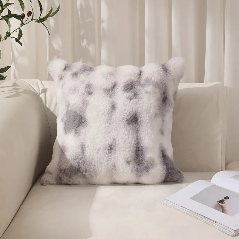 A fluffy faux rabbit fur decorative throw pillow with a gray and white mottled pattern resting on a light-colored sofa.