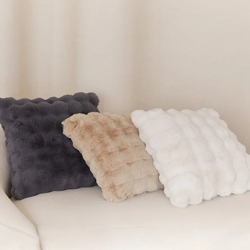 Three plush cushion covers in shades of gray, beige, and white resting on a light-colored sofa.
