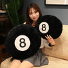 A person is holding a black 8 ball plush pillow with a white number 8 on it, sitting on a light-colored sofa.
