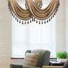 A beige window curtain valance with tassels hangs over a window in a bright room.