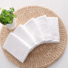 A stack of ten white cotton face towels resting on a woven mat next to a small potted plant.