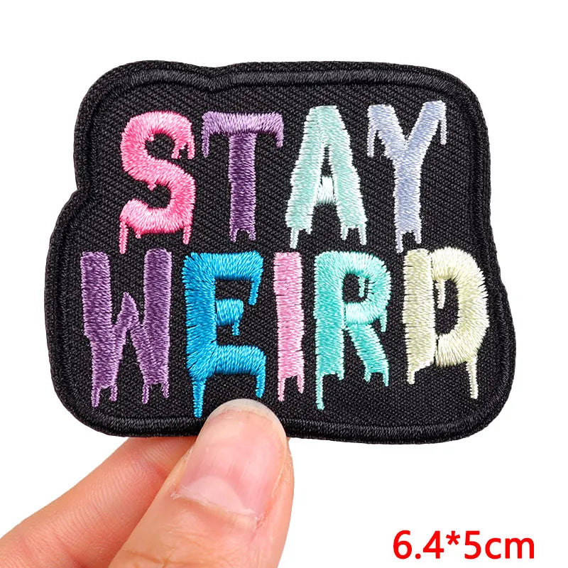 A black embroidered patch with the text STAY WEIRD in various pastel colors.