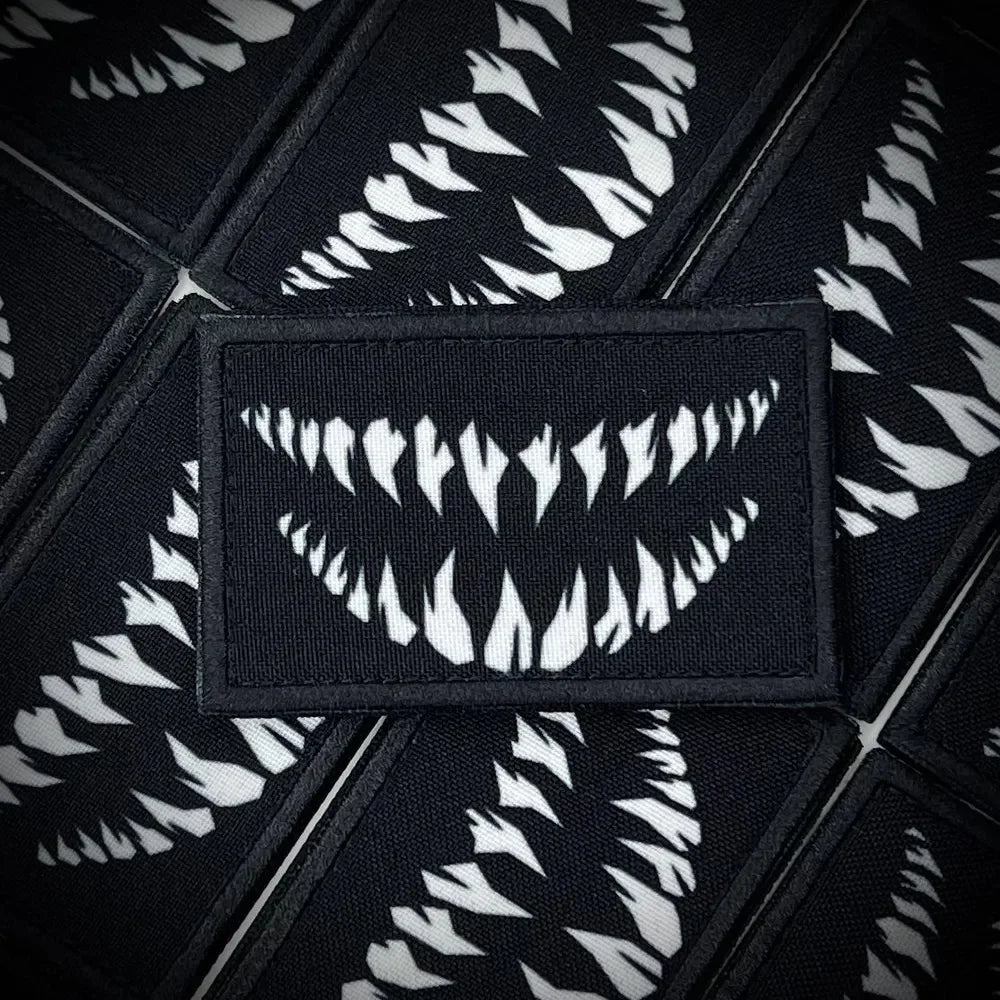 A close-up of a black tactical hook and loop emblem patch with white stylized teeth design.