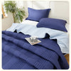 A navy blue quilted blanket and matching pillows are displayed on a bed with white linens.