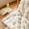 A fluffy cream faux fur throw blanket draped on a wooden floor next to a glass of white wine and an open book.