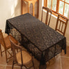 A dark lace tablecloth with floral embroidery is spread on a wooden dining table in a sunlit room.