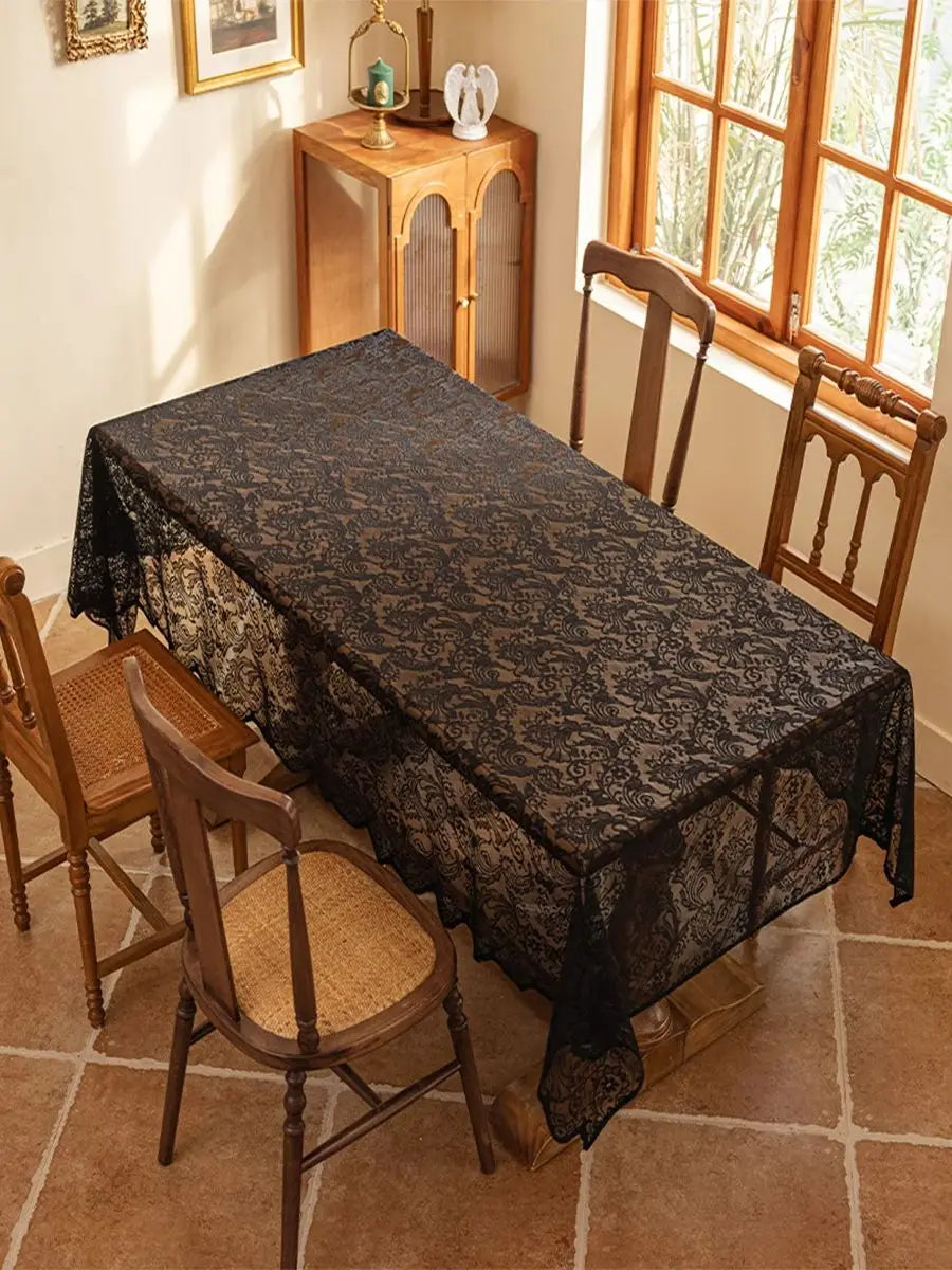 A dark lace tablecloth with floral embroidery is spread on a wooden dining table in a sunlit room.