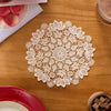 A white floral lace doily is centered on a wooden table next to a red plate and some fruit.