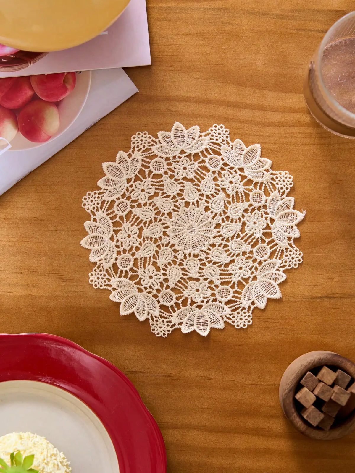 A white floral lace doily is centered on a wooden table next to a red plate and some fruit.