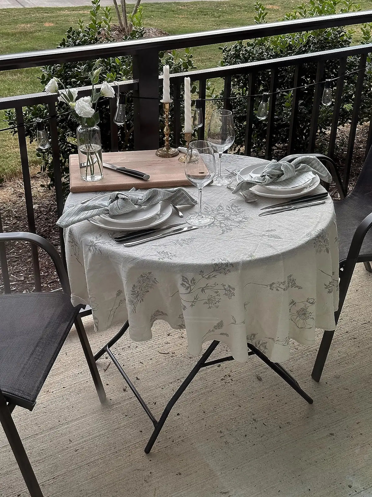 A round table set for dining with a botanical print linen tablecloth, glassware, and silverware on an outdoor patio.