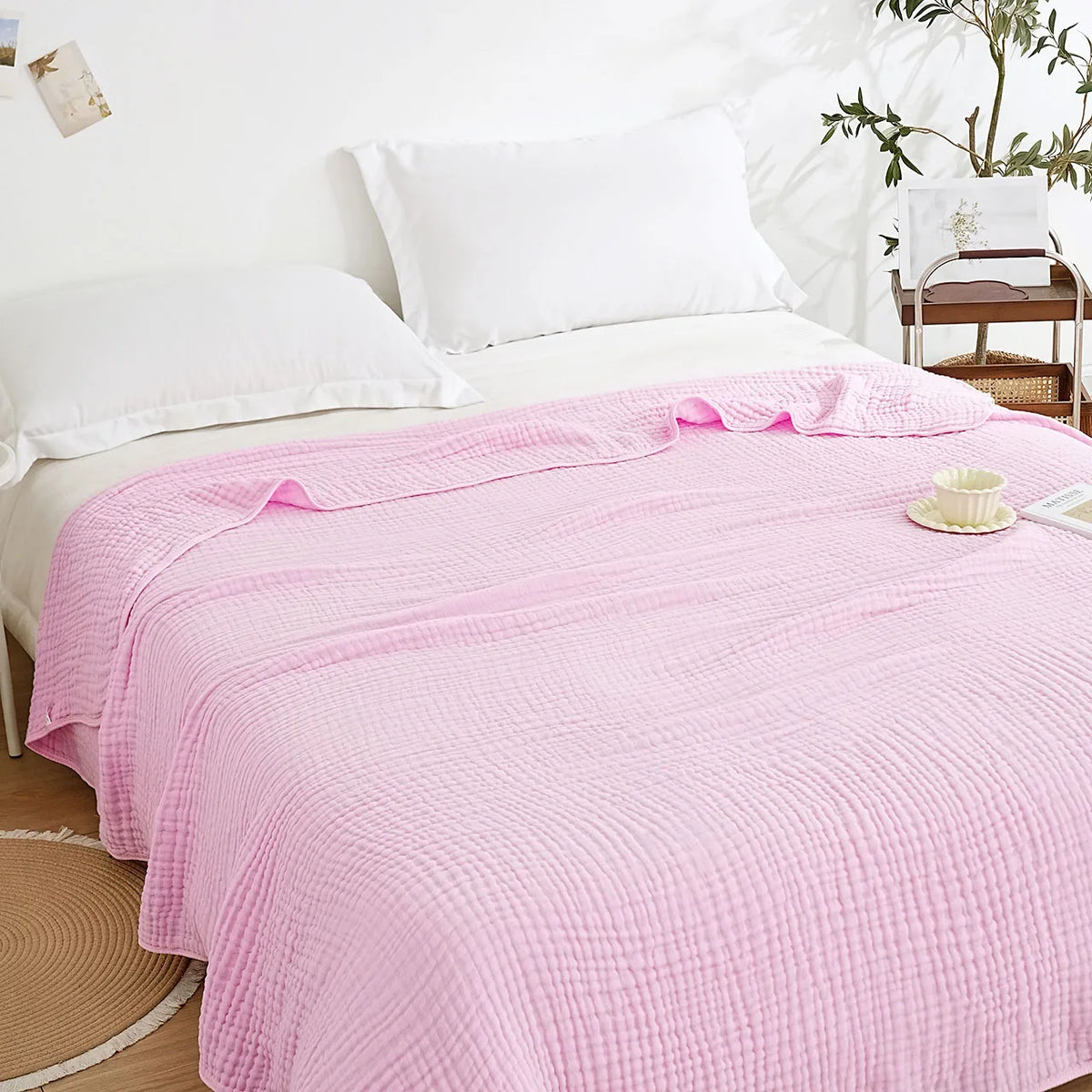 A pink quilted muslin blanket is draped over a white bed with white pillows.