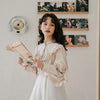 A young woman wearing a cream-colored retro lace long sleeve blouse and white skirt, holding a newspaper.