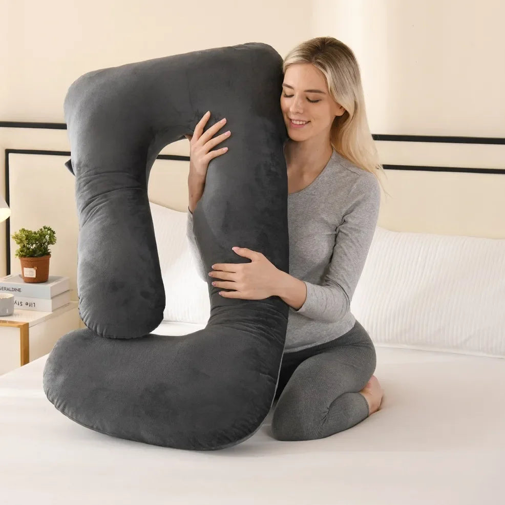 A woman is lying down, embracing a gray ergonomic J-shaped maternity pillow on a white bed.