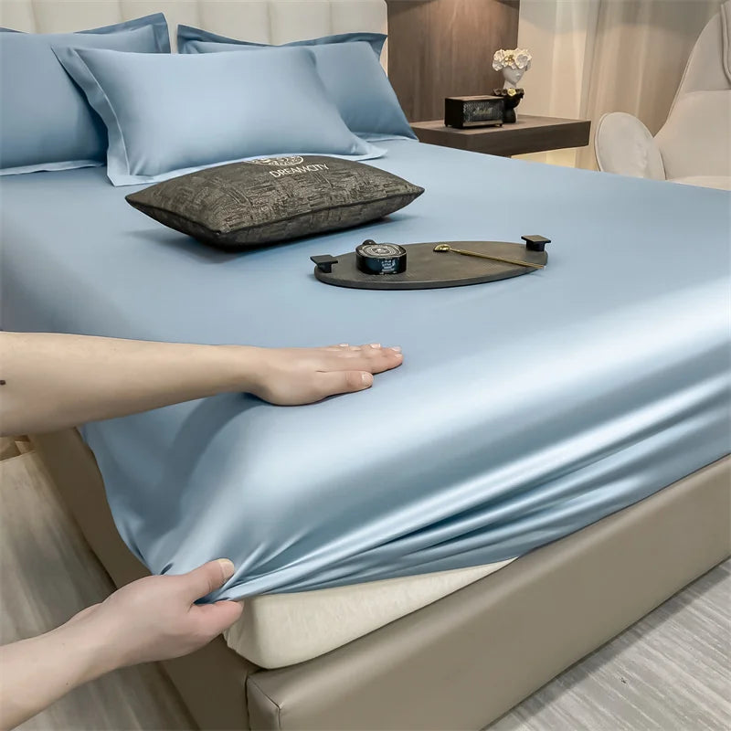 A light blue fitted bed sheet is draped over a bed, with pillows and a small tray on the surface.