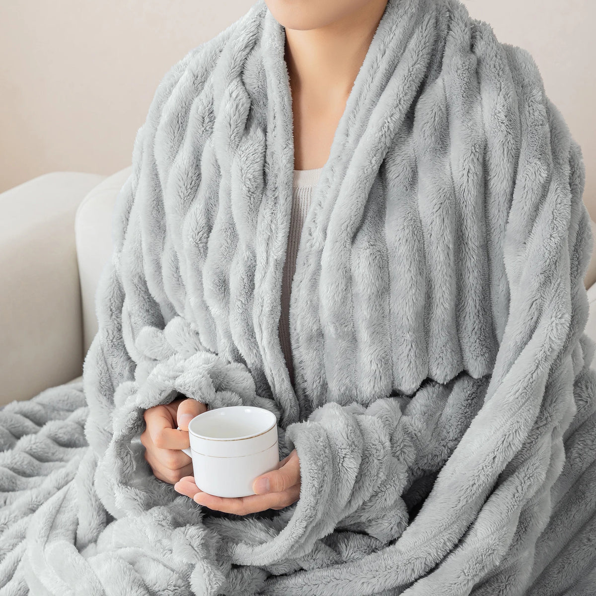 A person is wrapped in a thick, gray soft flannel throw blanket while holding a white mug.