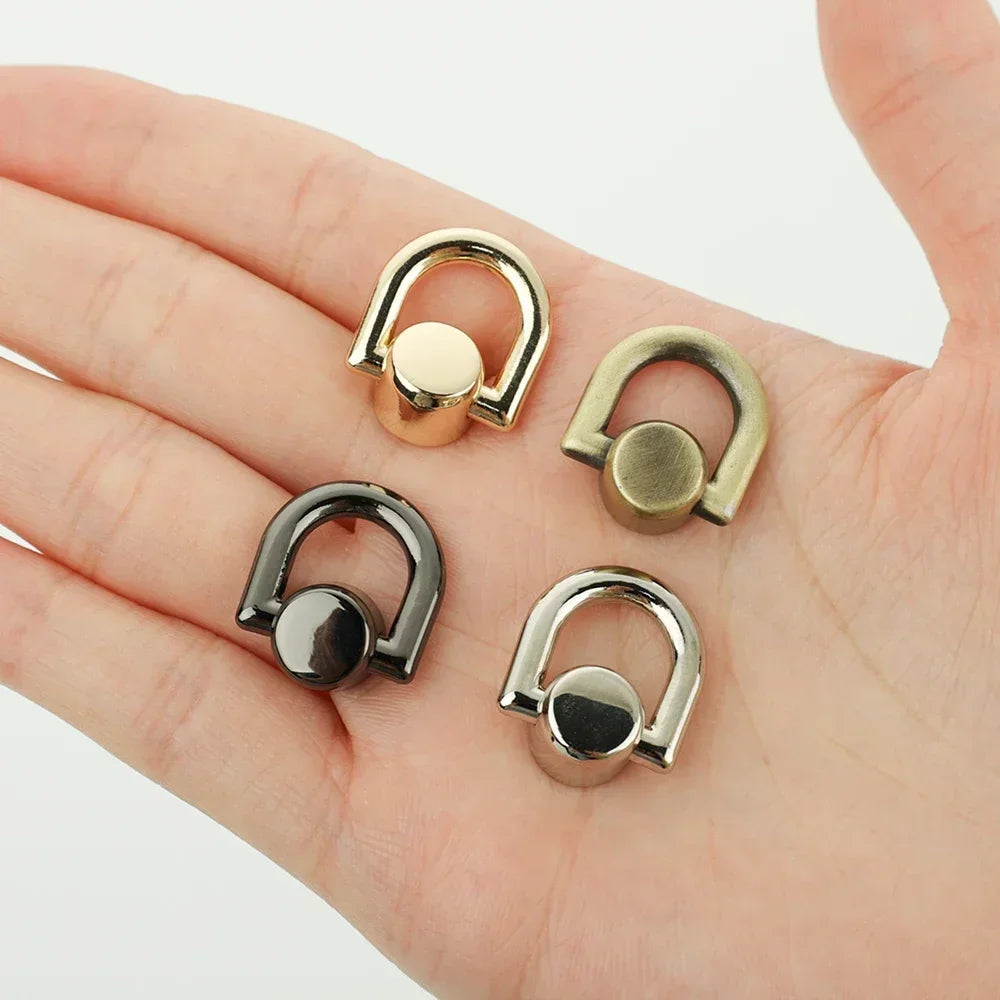 A hand holds several metal D-shaped ring bag handles with screws in various gold and silver tones.