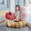 A woman is sitting on the floor surrounded by Yayoi Kusama pumpkin plush cushions in orange, yellow, and white polka dots.