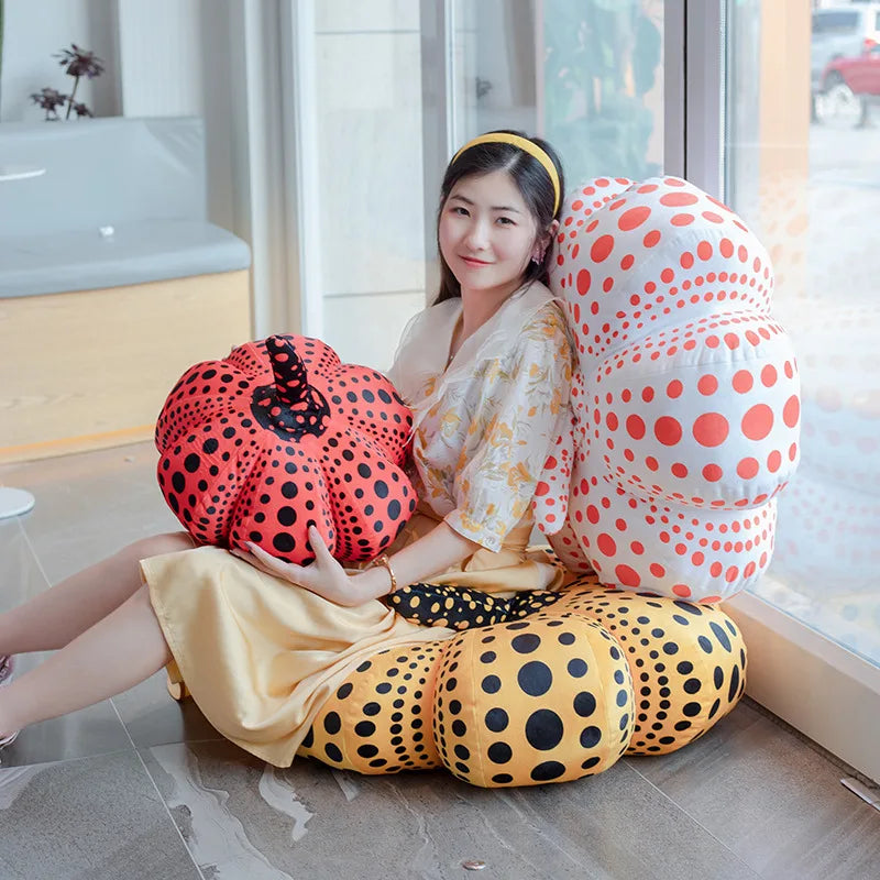 A woman is sitting on the floor surrounded by Yayoi Kusama pumpkin plush cushions in orange, yellow, and white polka dots.