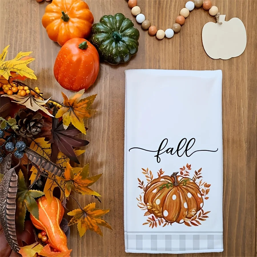 A white kitchen towel with an orange pumpkin print and a decorative fall arrangement.
