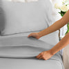 A hand is smoothing a gray microfiber bed sheet on a bed with a tufted headboard.