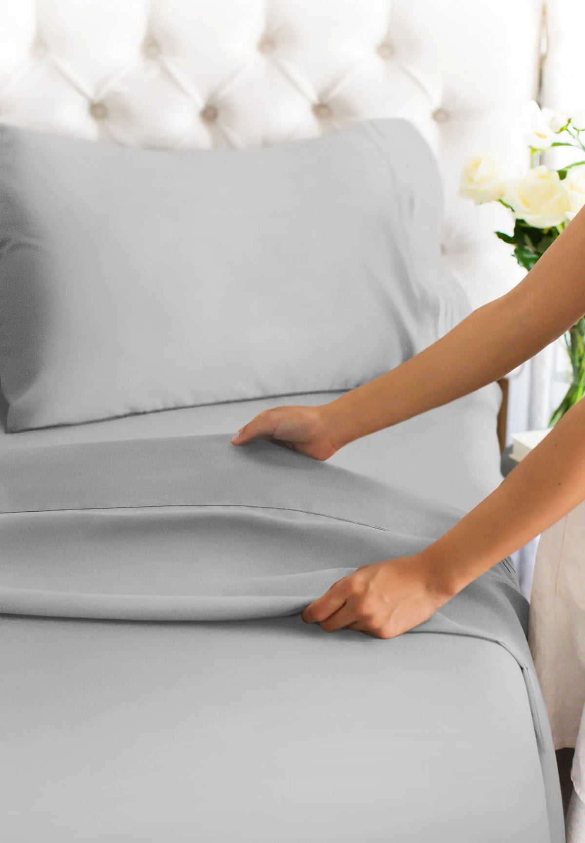 A hand is smoothing a gray microfiber bed sheet on a bed with a tufted headboard.