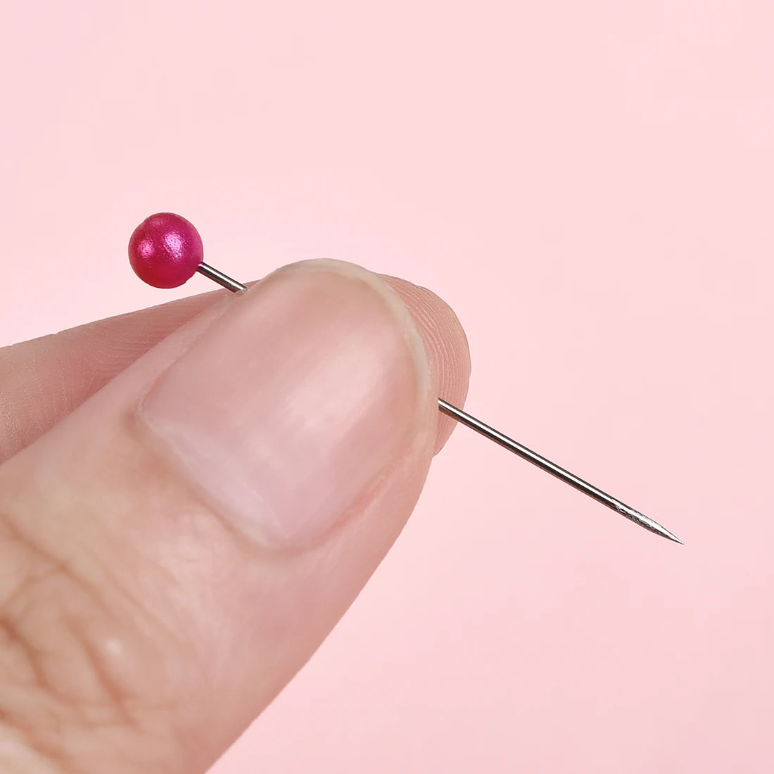 A hand holds a pink pearl head sewing pin against a light pink background.