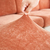 A hand is adjusting a textured, rust-colored chenille sofa cushion cover on an orange upholstered sofa.