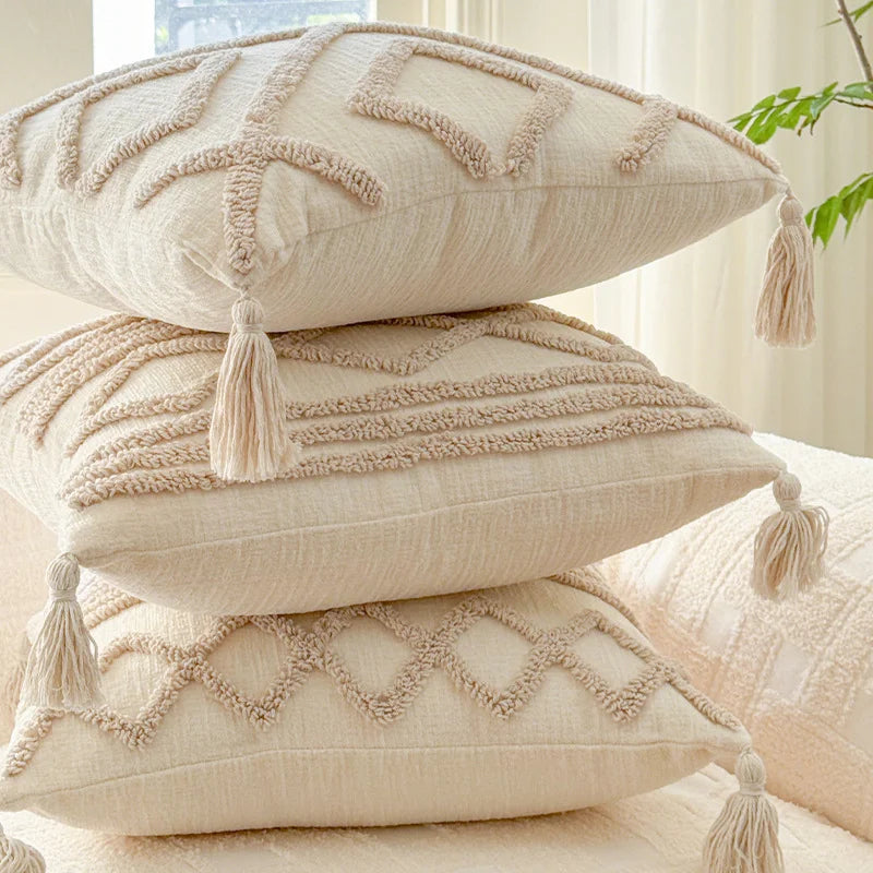 Three stacked beige knit pillow covers with fringed edges on a light-colored surface.