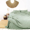 A luxury French linen duvet cover in sage green draped over a bed with a natural fiber wall hanging.