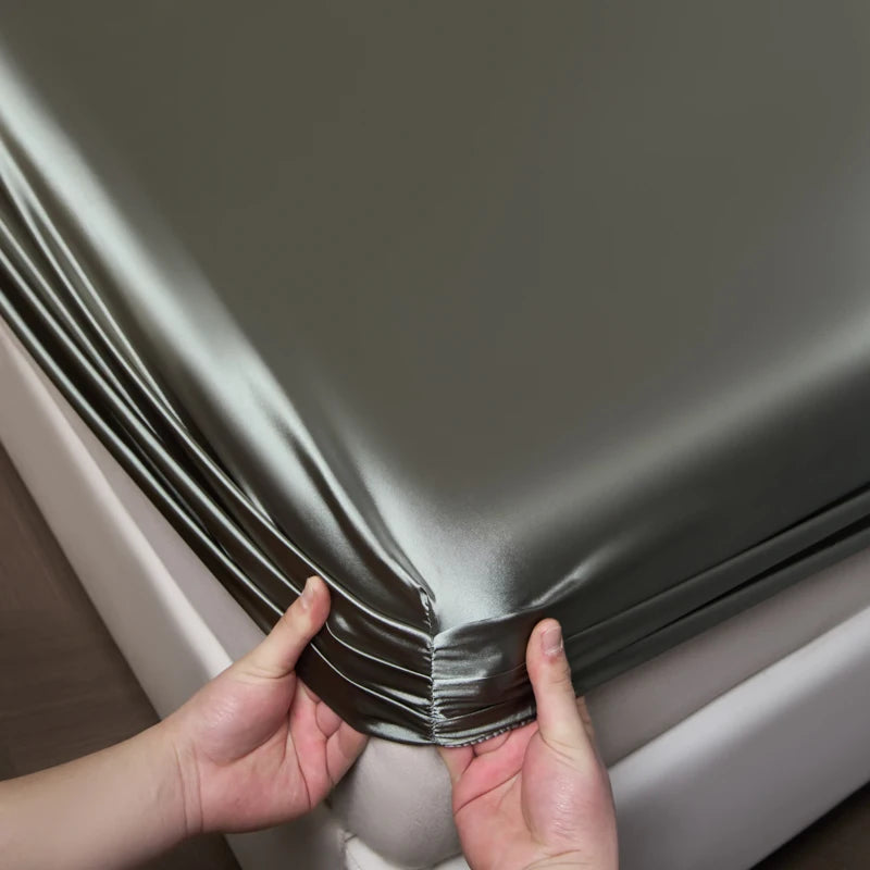 Hands are folding a silky gray satin fitted sheet over a white mattress.