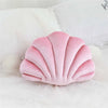 A pink velvet stuffed sofa cushion shaped like a decorative seashell resting on white fabric.