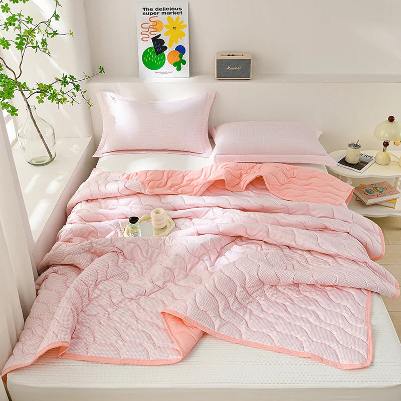 A pink quilted lightweight cooling summer quilt set displayed on a white bed with a green plant and a small box.