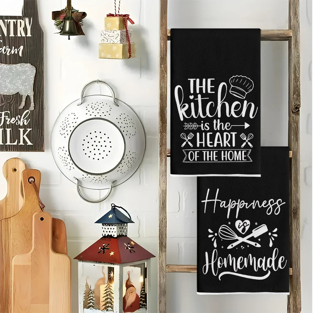 A set of two black kitchen towels featuring designs for the kitchen, heart of the home, and happiness homemade