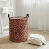 A foldable leopard print laundry basket with a black handle sitting on a beige rug next to a white window.