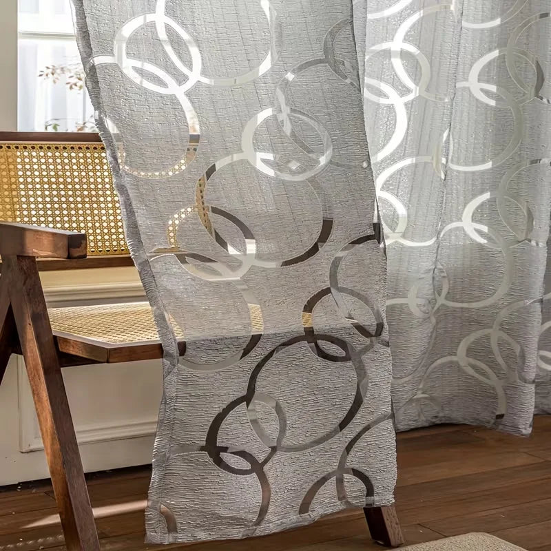 A sheer white curtain with geometric circular patterns in brown embroidery hanging indoors.
