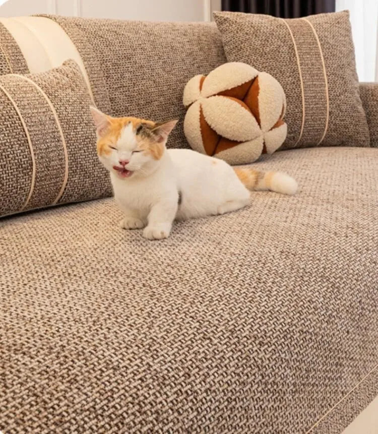 A small orange and white cat is lying on a textured gray sofa cushion surrounded by patterned pillows.