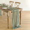 A rustic gauze table runner in sage green decorates a wooden table set for dining.