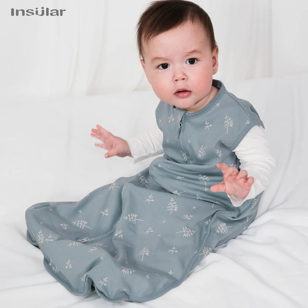 A baby wearing a light blue cotton sleep sack with a white pattern is lying on a white surface.