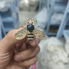A hand holds a gold-toned crystal bee brooch with a dark blue pearl accent.