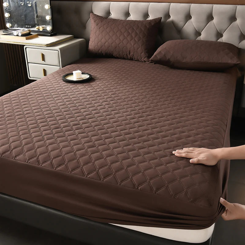A dark brown quilted mattress protector is shown on a bed, next to a bedside table.