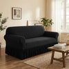 A black dustproof stretch sofa slipcover placed on a wooden floor with a rug and natural light.