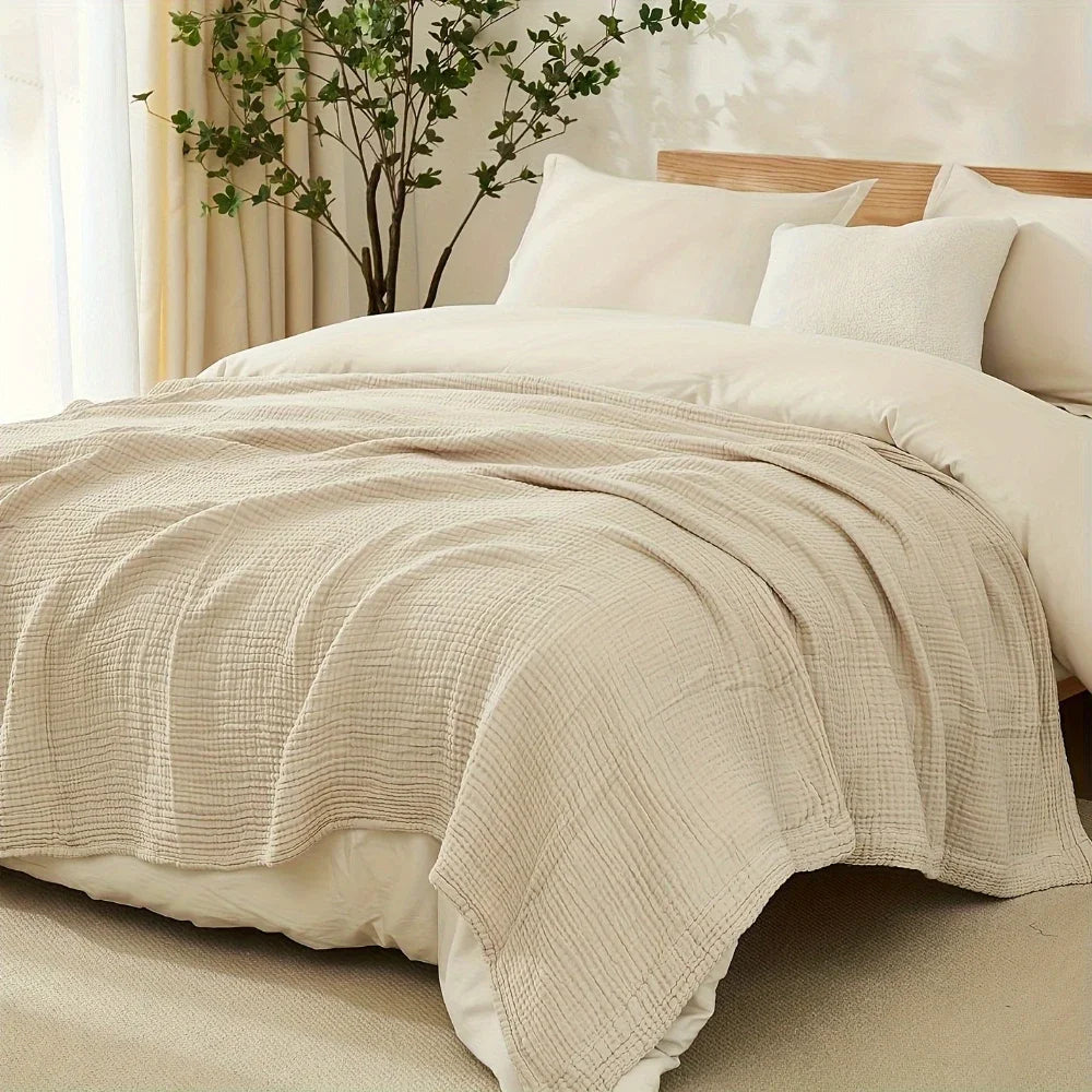 A beige cotton gauze blanket draped over a bed, next to a wooden headboard and green foliage.