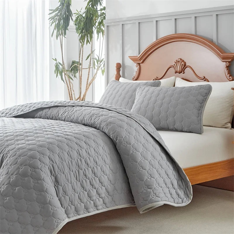 A gray quilted bedspread set is draped over a white bed with a wooden headboard and sheer curtains.