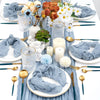 A rustic boho table setting featuring a blue cheesecloth runner, gold cutlery, and white floral arrangements.
