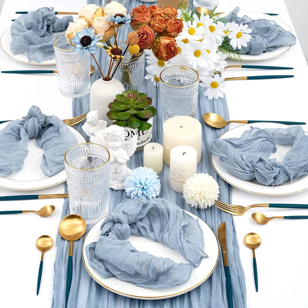 A rustic boho table setting featuring a blue cheesecloth runner, gold cutlery, and white floral arrangements.